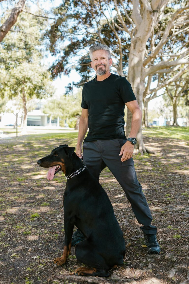 A man poses with a Doberman in a green sunny park, embodying companionship.